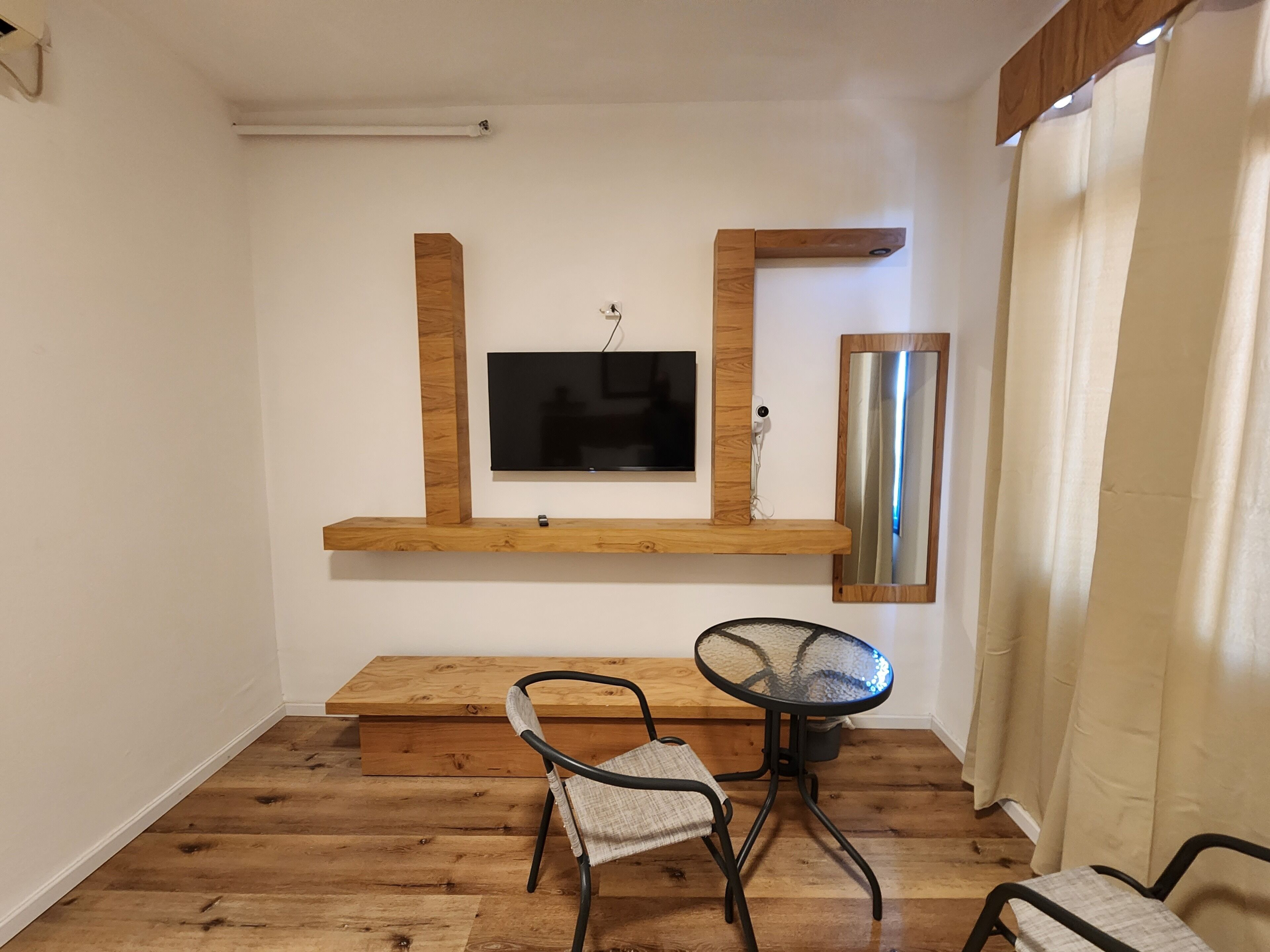 Una stanza con pavimento in legno, una TV montata su una mensola di legno, un tavolino in vetro con due sedie e uno specchio a parete.
