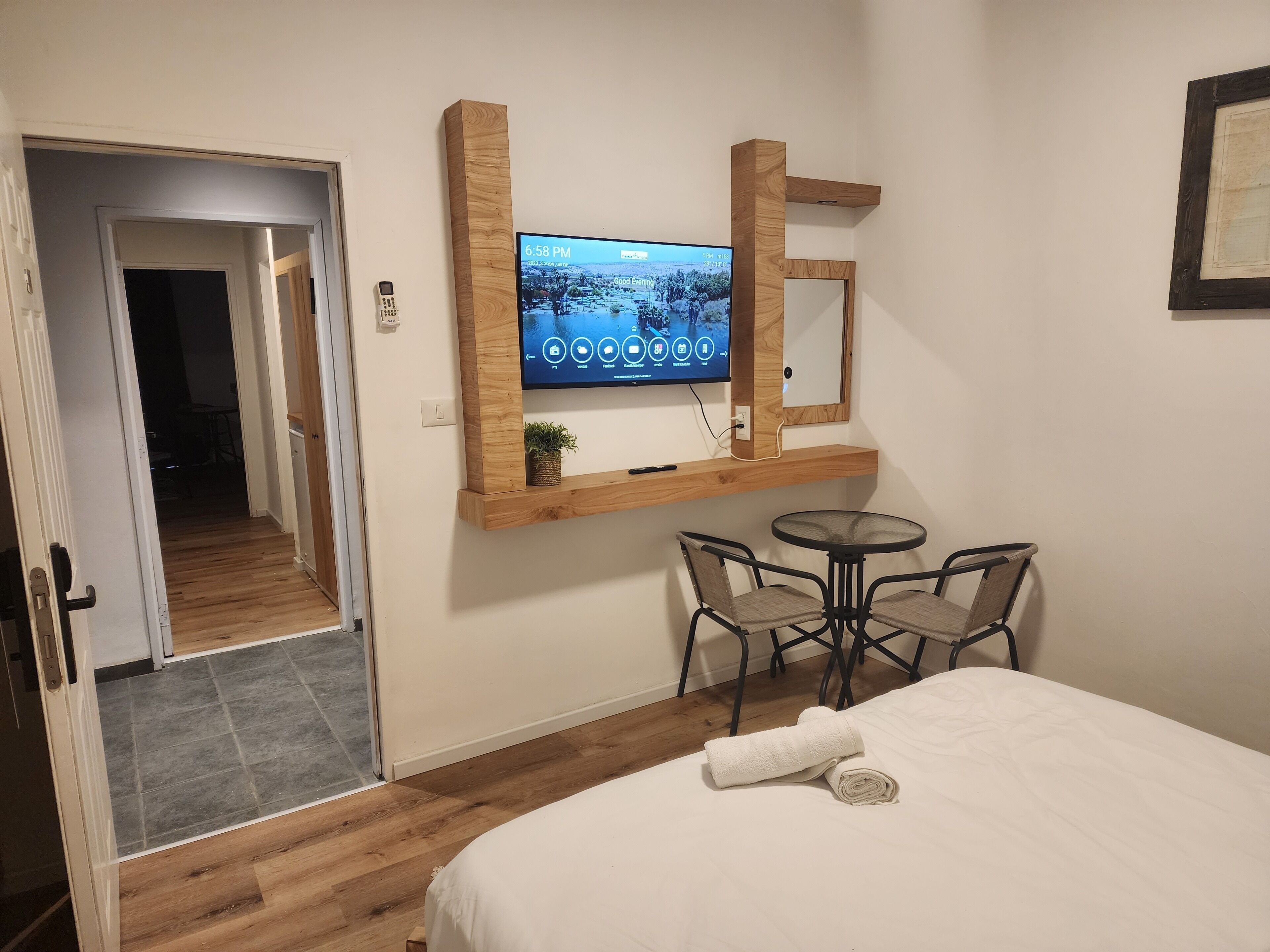 A hotel room with a bed, a TV mounted on a wooden shelf, a small table with chairs, and a door leading to another room.
