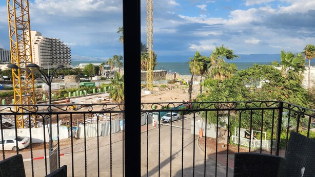 A balcony with a view of a construction site, palm trees, and a building with a sign that reads "tenoardo." - Tiberia hotel (Tiberias)