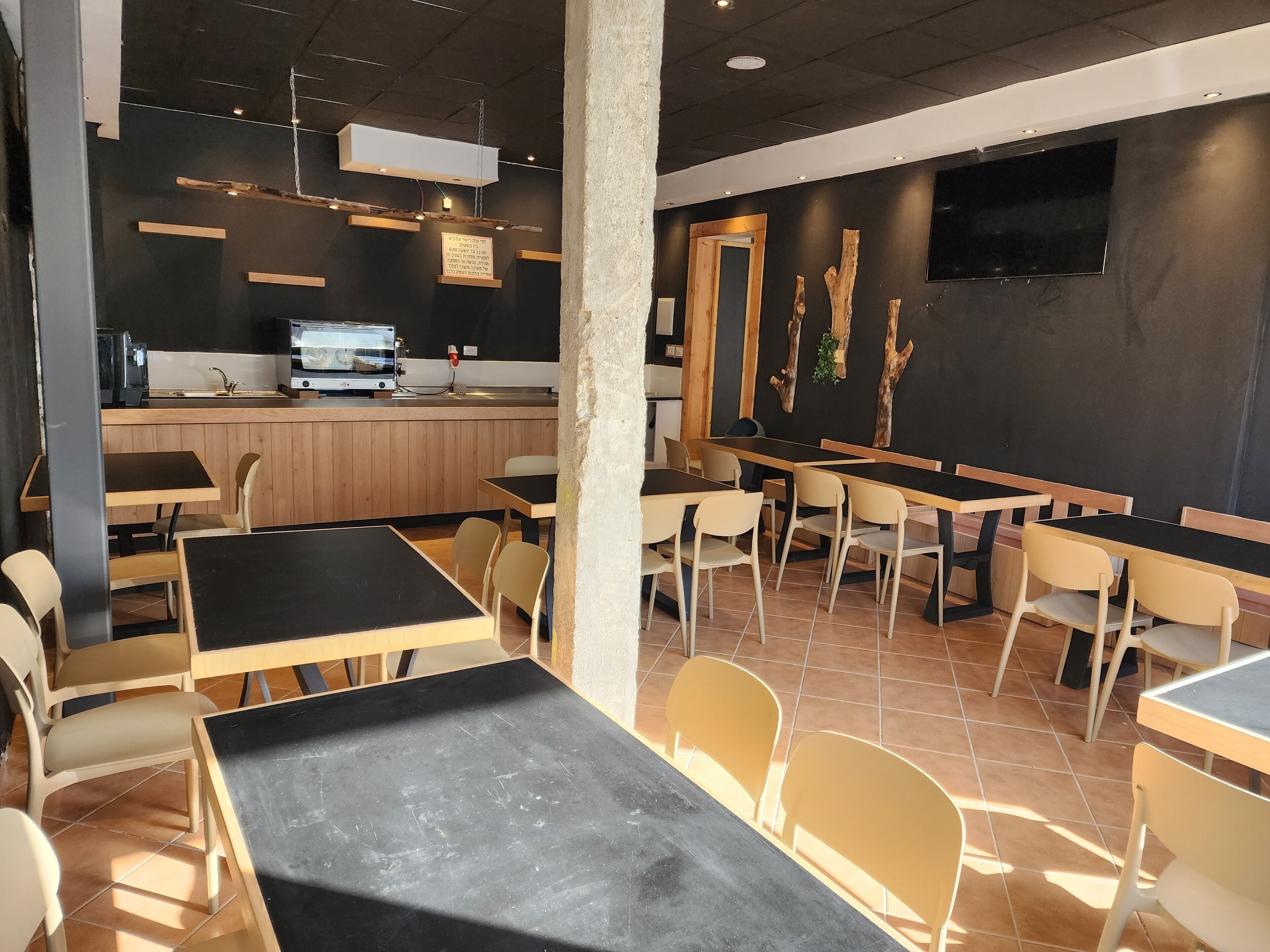 Un restaurant au décor moderne, avec un mélange de tables et de chaises en bois et en noir, une cuisine ouverte et un téléviseur à écran plat fixé au mur.