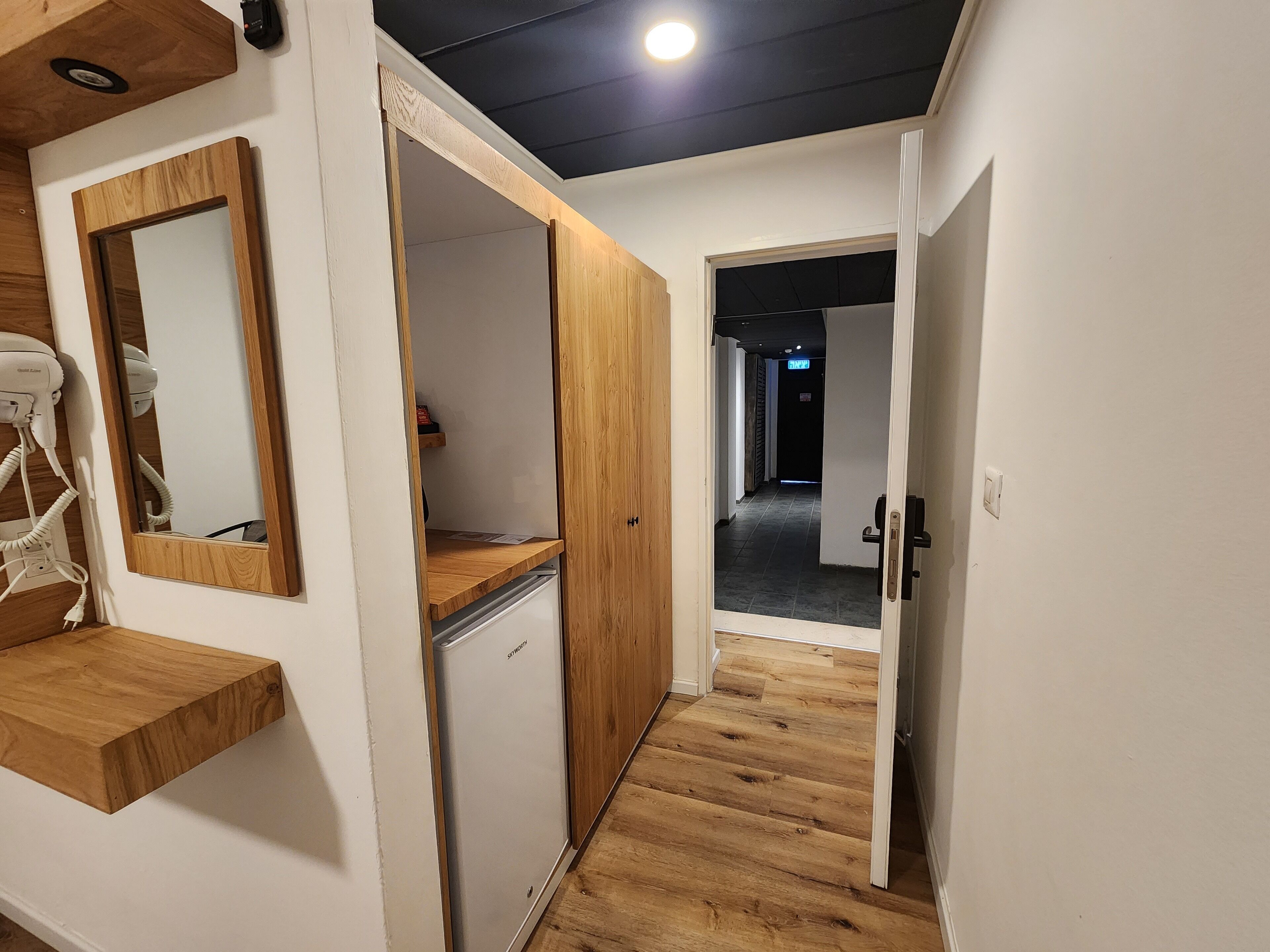 Un couloir avec un plancher en bois, un mur blanc et un miroir. On y trouve un petit appareil électroménager blanc, qui pourrait être un réfrigérateur, ainsi qu’une étagère en bois.
