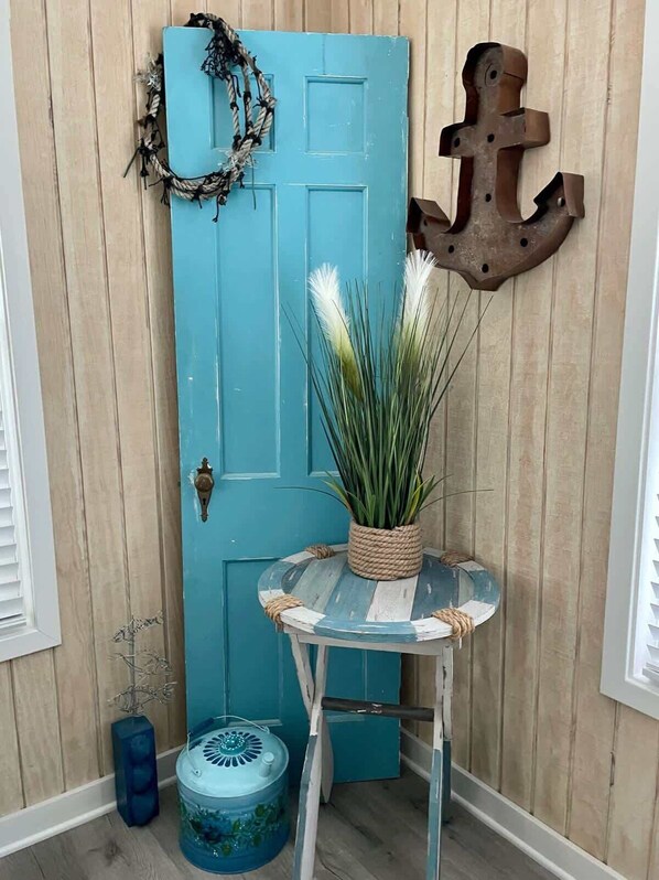 Une porte bleue ornée d’une guirlande, un pot de fleurs, une table ronde, une ancre décorative et un vase bleu.