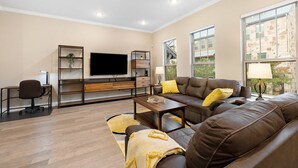 A modern living room with a brown leather sofa, a flat-screen TV, wooden shelves, and a desk with a chair. - Your Home Away From Home in the Texas Hill Country, Georgetown TX (Georgetown)