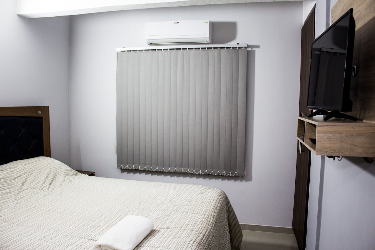 A hotel room with a bed, a TV, a window with blinds, and an air conditioning unit.