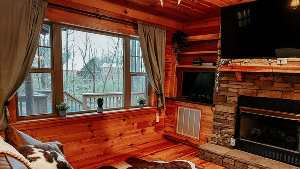 L’intérieur chaleureux d’un chalet avec des murs en bois, une cheminée en pierre, un téléviseur et une grande fenêtre agrémentée de rideaux.