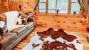 Un intérieur de chalet chaleureux, avec un revêtement mural en bois, un canapé recouvert d’une couverture, un tapis en peau de vache et une fenêtre agrémentée de rideaux.