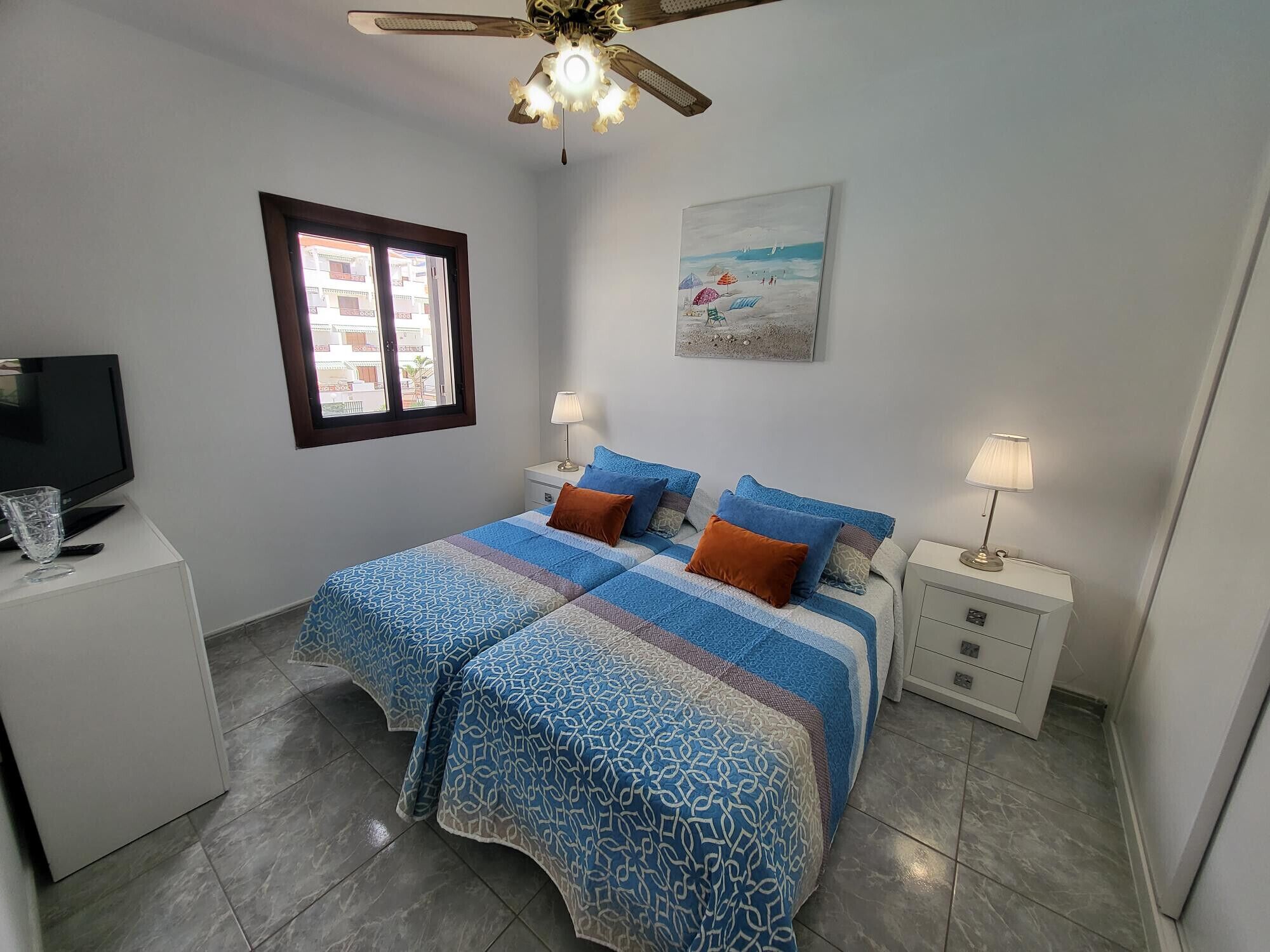 A bedroom with a bed, two bedside tables, a ceiling fan, a window, and a painting on the wall.