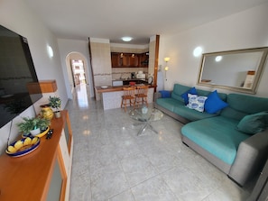 A modern living room with a teal sofa, a glass coffee table, and a kitchen area with wooden cabinets and a dining table.