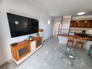A modern living room with a flat-screen TV, wooden furniture, a glass coffee table, and a kitchen area with wooden cabinets and a tiled backsplash.