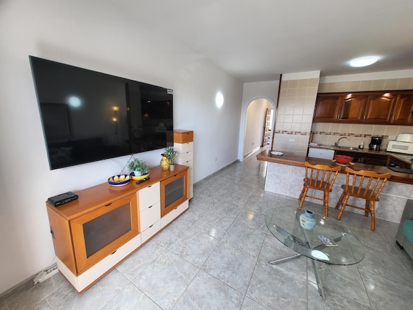 A modern living room with a flat-screen TV, wooden furniture, a glass coffee table, and a kitchen area with wooden cabinets and a tiled backsplash.