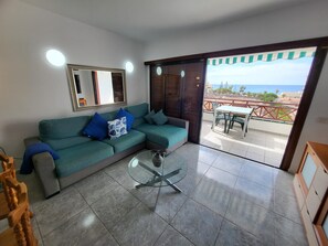 A living room with a green sofa, a glass coffee table, a wooden chair, a dining table with chairs, and a view of the sea through a balcony.