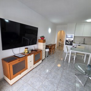 A modern living room with a flat-screen TV, a dining area with a glass table, and a kitchen with white cabinets and a microwave. - Victoria Court II (Los Cristianos)
