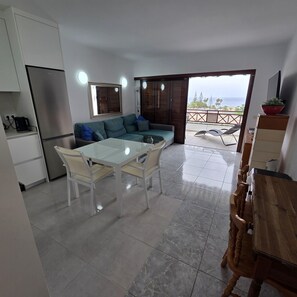 A modern kitchen with a refrigerator, a dining table with chairs, and a sofa. There is a balcony with a view of the sea. - Victoria Court II (Los Cristianos)