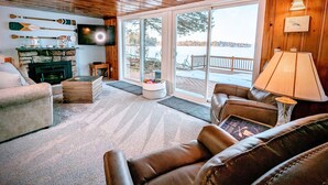 A cozy living room with a stone fireplace, leather sofas, a television, and a view of a snowy landscape through large sliding glass doors.