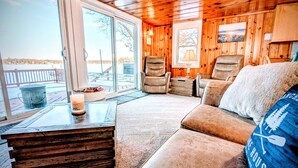 A cozy cabin interior with a large window overlooking a deck, a leather sofa, and a rustic wooden ceiling.