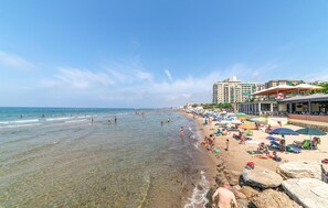 Uma praia com águas cristalinas e rasas, faixa de areia e fileira de edifícios com varandas voltadas para a praia.