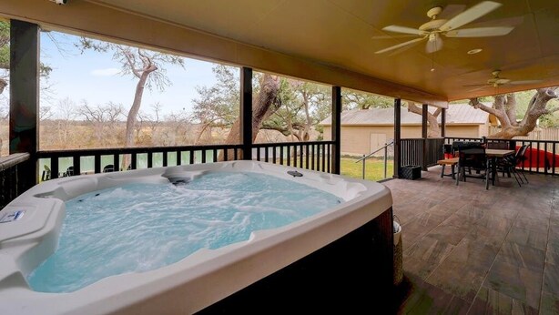 A hot tub in an outdoor patio area with a ceiling fan, a table and chairs, and a view of trees and a house in the background. - Longhorn River Ranch (Seguin)