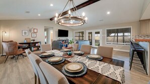 A dining area with a wooden table set for six, surrounded by chairs. A chandelier hangs above the table, and there is a television mounted on the wall. - Longhorn River Ranch (Seguin)