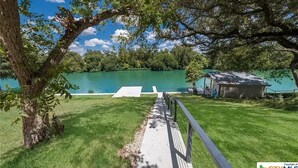 A grassy area with a pathway leading to a body of water, surrounded by trees and a small structure. - Longhorn River Ranch (Seguin)