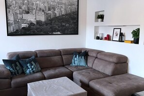 A living room with a brown sectional sofa, a small beige ottoman, and a cityscape painting on the wall. There are shelves with decorative items and a potted plant.