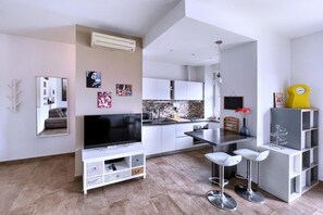 Una cocina moderna con armarios blancos, salpicadero de azulejos y una isla central. Hay un comedor con taburetes de bar blancos y una mesita.