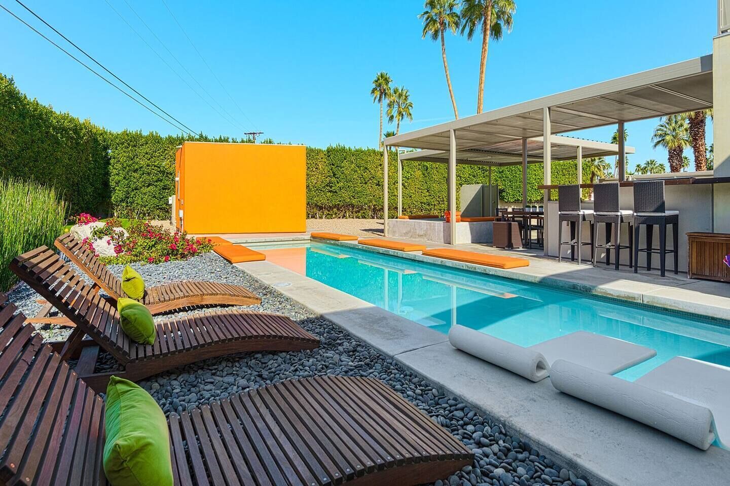 A modern outdoor pool area with wooden loungers, a covered seating area, and a large orange wall.