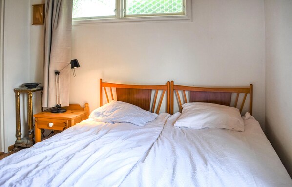 A single bed with a wooden headboard in a room with white walls, a small bedside table, and a window with a view of greenery.