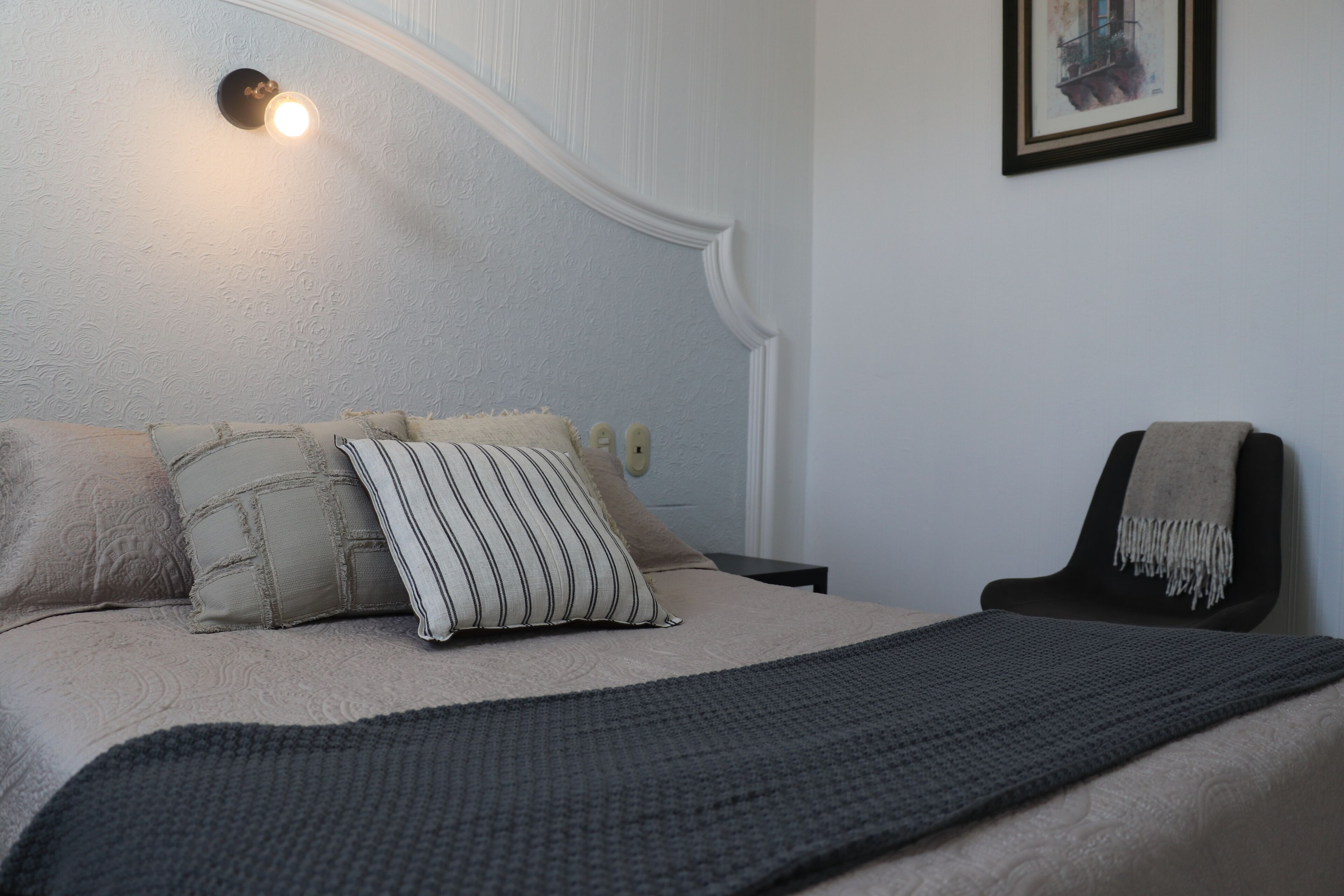 A bedroom with a bed, pillows, a blanket, a chair, a wall-mounted light, and a framed picture.