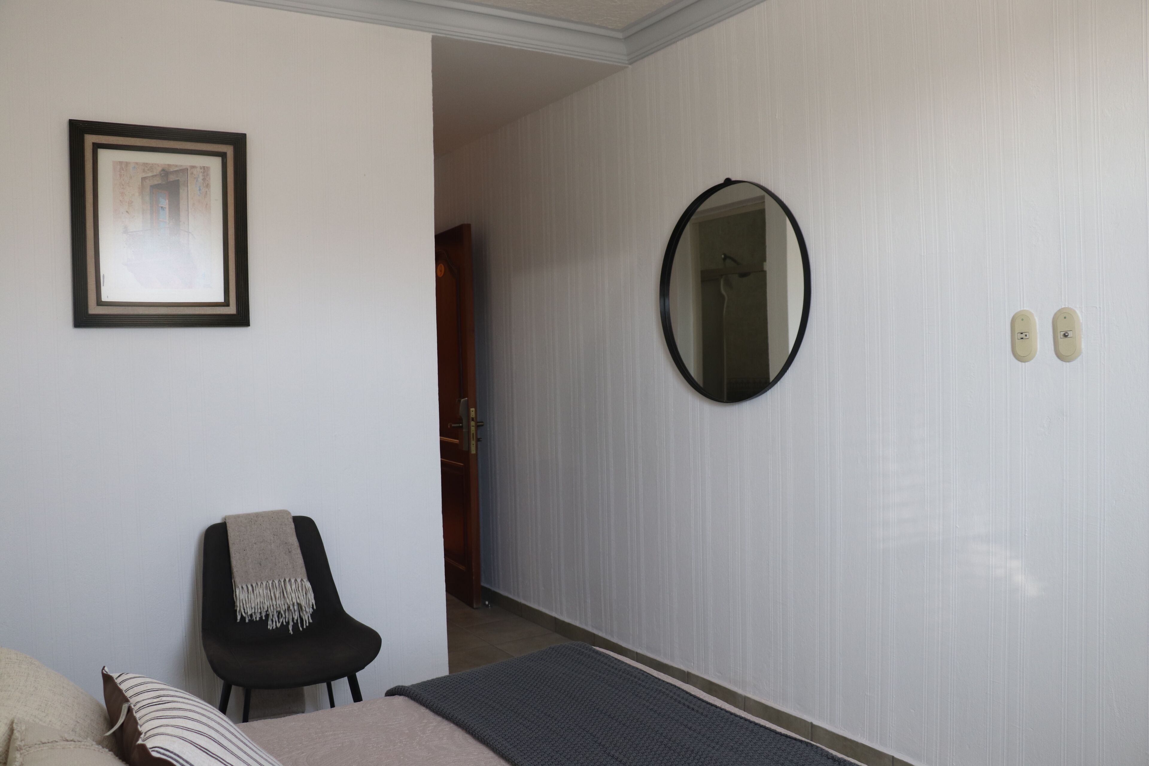 Une chambre d’hôtel avec un lit, une chaise recouverte d’une couverture, un tableau encadré au mur, un miroir et une porte donnant sur une autre pièce.