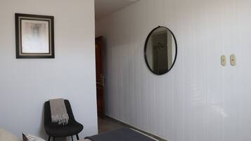 Une chambre d’hôtel avec un lit, une chaise recouverte d’une couverture, un tableau encadré au mur, un miroir et une porte donnant sur une autre pièce.
