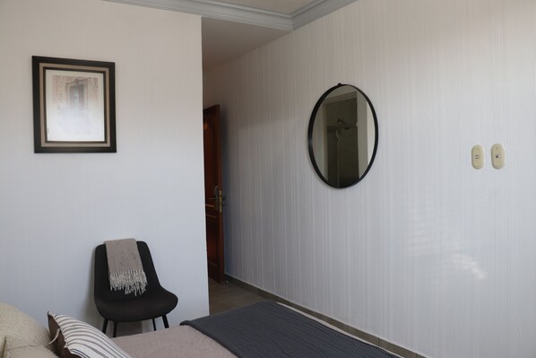 Habitación de hotel con una cama, una silla con una manta, un cuadro enmarcado en la pared, un espejo y una puerta que da a otra habitación.