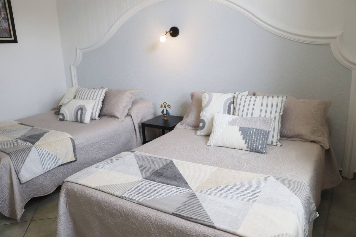Une chambre d’hôtel avec deux lits, chacun recouvert d’une couvre-lit à motifs géométriques et agrémenté d’oreillers assortis. Une table de chevet avec une lampe se trouve entre les deux lits.