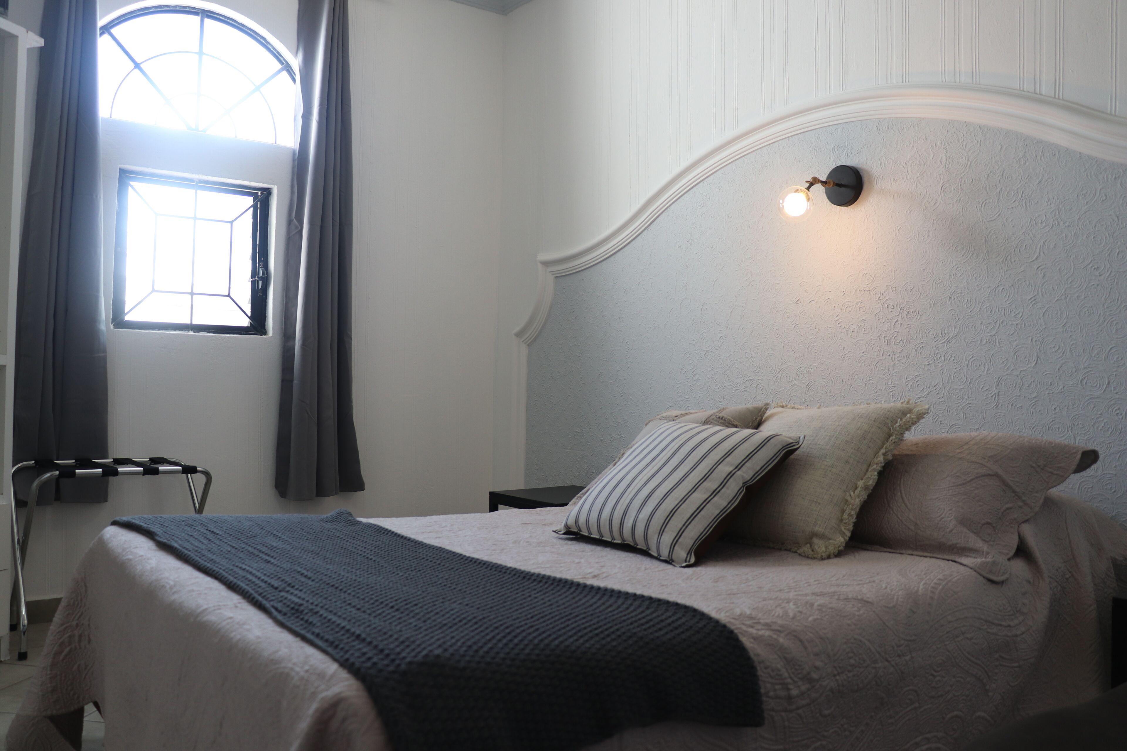 A neatly made bed with a striped pillow and a blanket, a bedside table, a wall-mounted light, and a window with curtains.