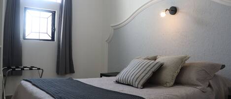 A neatly made bed with a striped pillow and a blanket, a bedside table, a wall-mounted light, and a window with curtains.