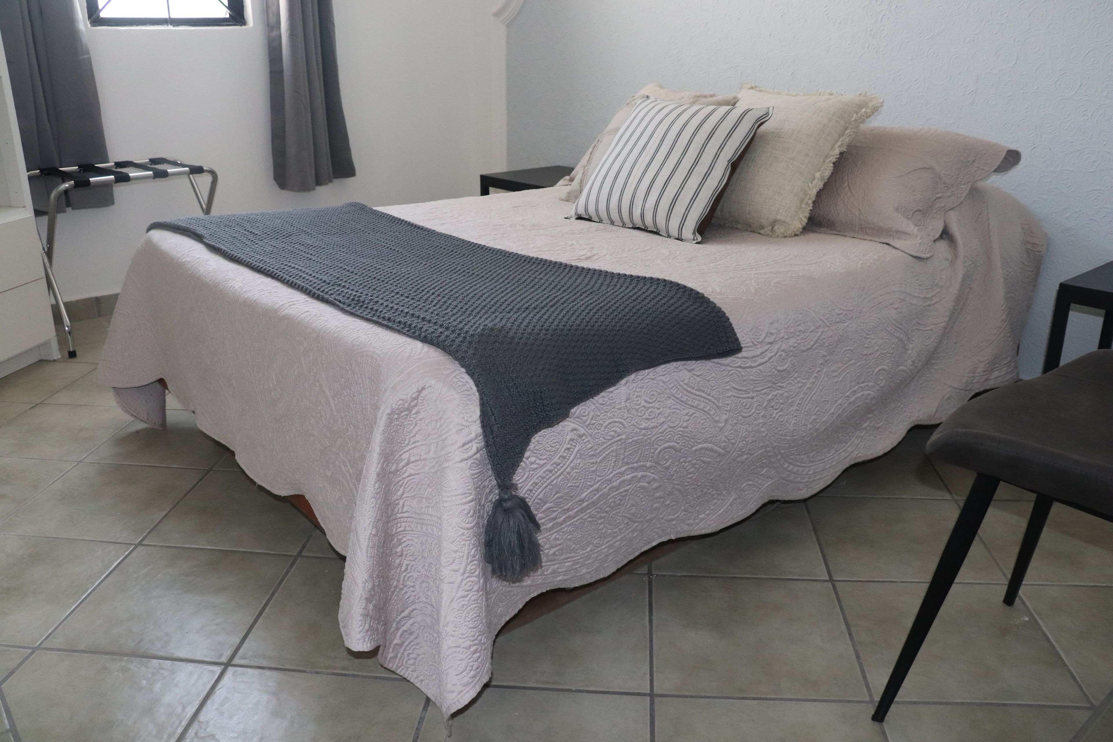 A neatly made bed with a grey blanket and a tassel, a bedside table, a chair, and a window with curtains.