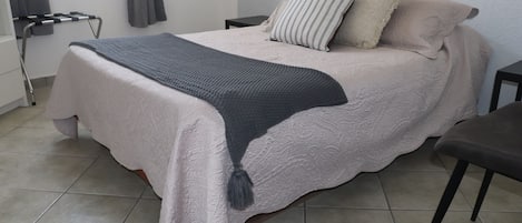 A neatly made bed with a grey blanket and a tassel, a bedside table, a chair, and a window with curtains.