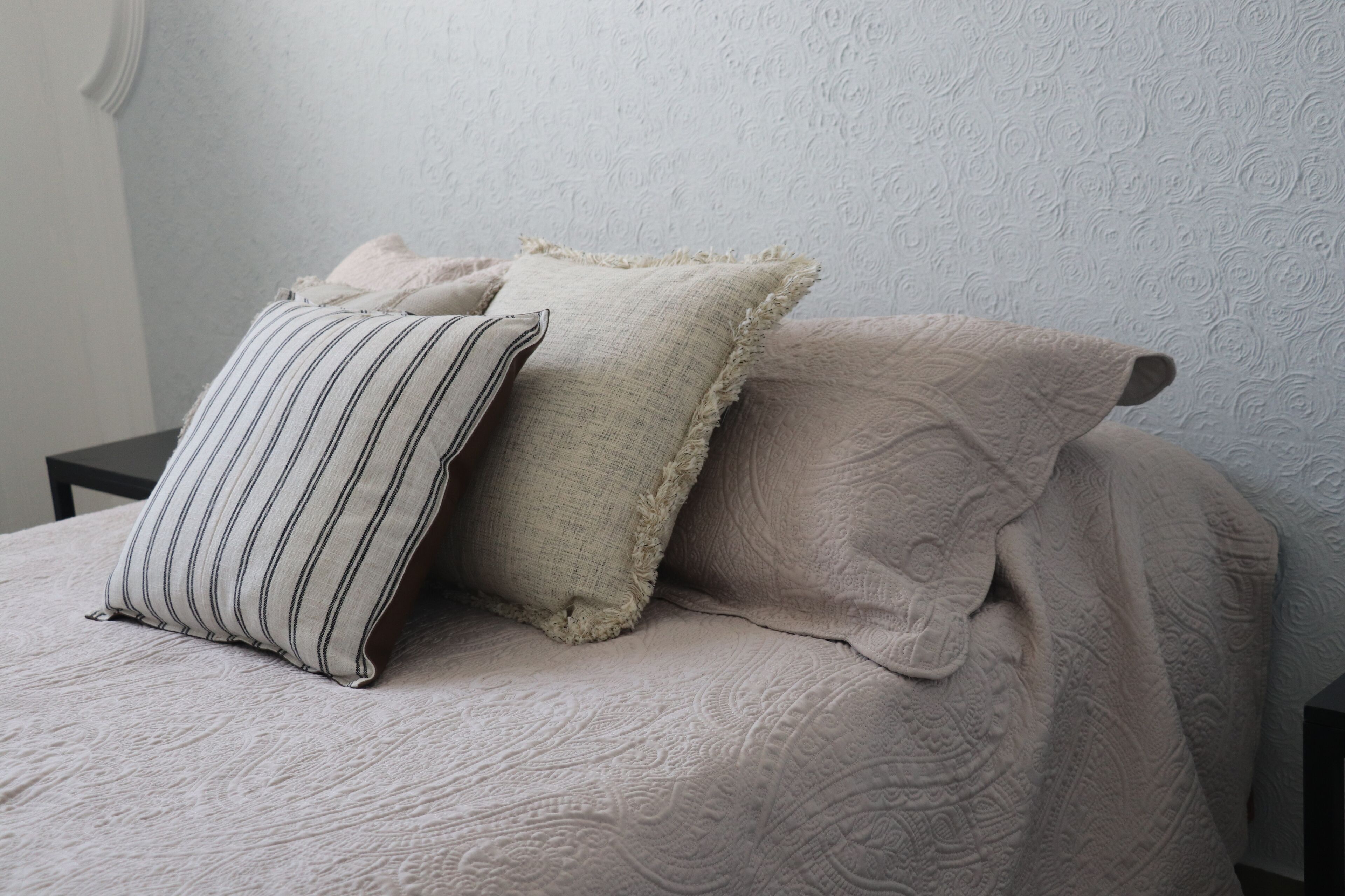 A bed with a textured beige bedspread and three pillows, one striped, one with a rough texture, and one with a floral pattern.