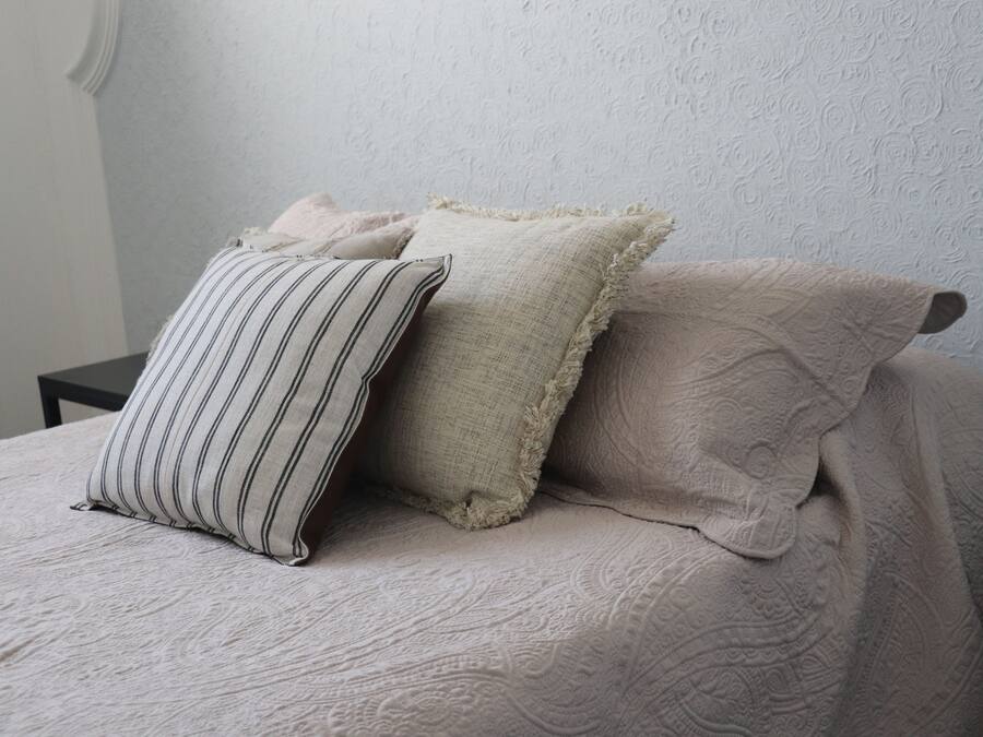 Een bed met een geribbelde beige sprei en drie kussens, waarvan één gestreept, één met een ruwe textuur, en één met een bloemenprint.