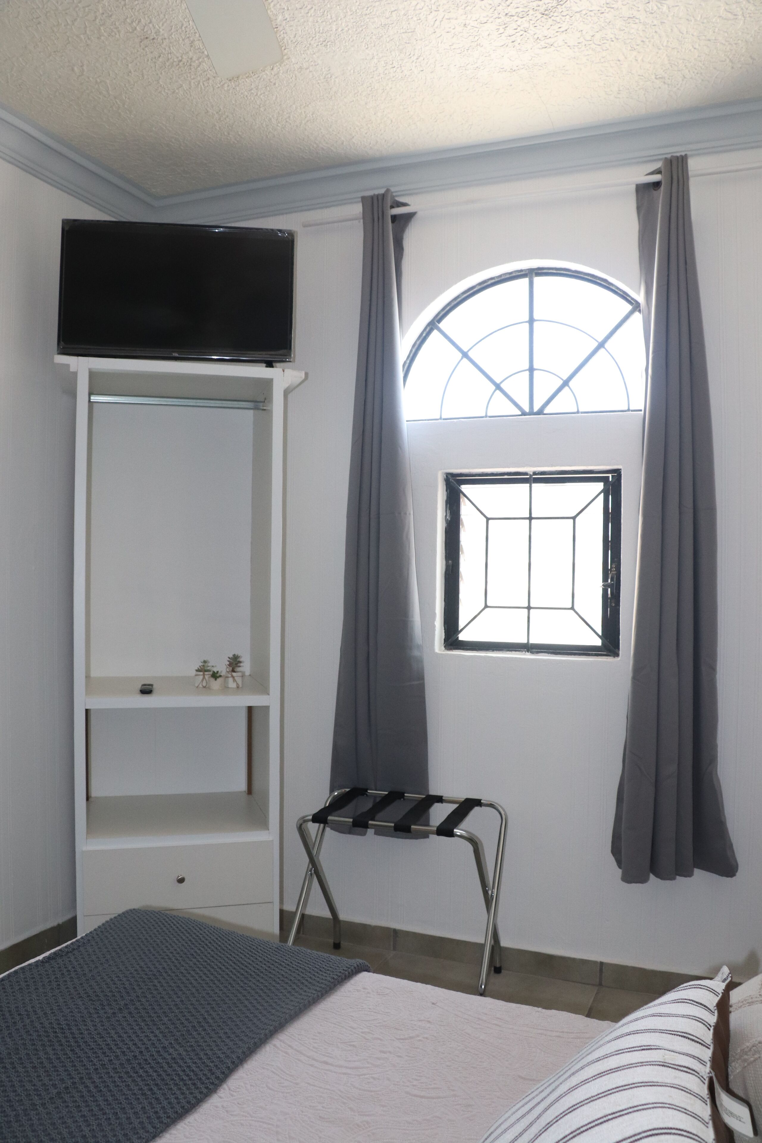 Une chambre à coucher comprenant un lit, un téléviseur fixé au mur, une fenêtre avec des rideaux et une petite étagère ornée d’objets décoratifs.