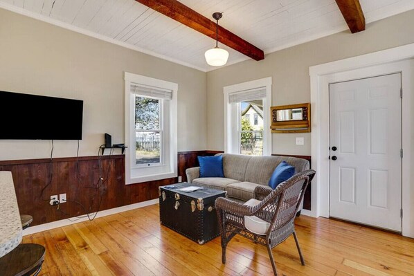 A living room with a wooden floor, a sofa with blue cushions, a wicker chair, a small table, and a white door.