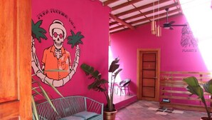 A room with a vibrant pink wall, a metal bench, a wooden door, and a ceiling fan. - Planet B Hostel (Quepos)