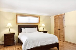 Une chambre à coucher comprenant un lit, deux tables de chevet avec des lampes, une tête de lit en bois, une fenêtre avec des stores et une armoire en bois.