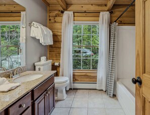 Une salle de bain avec un plafond en bois, un lavabo doté d’un plan de travail en granit, des toilettes et une fenêtre avec des rideaux.
