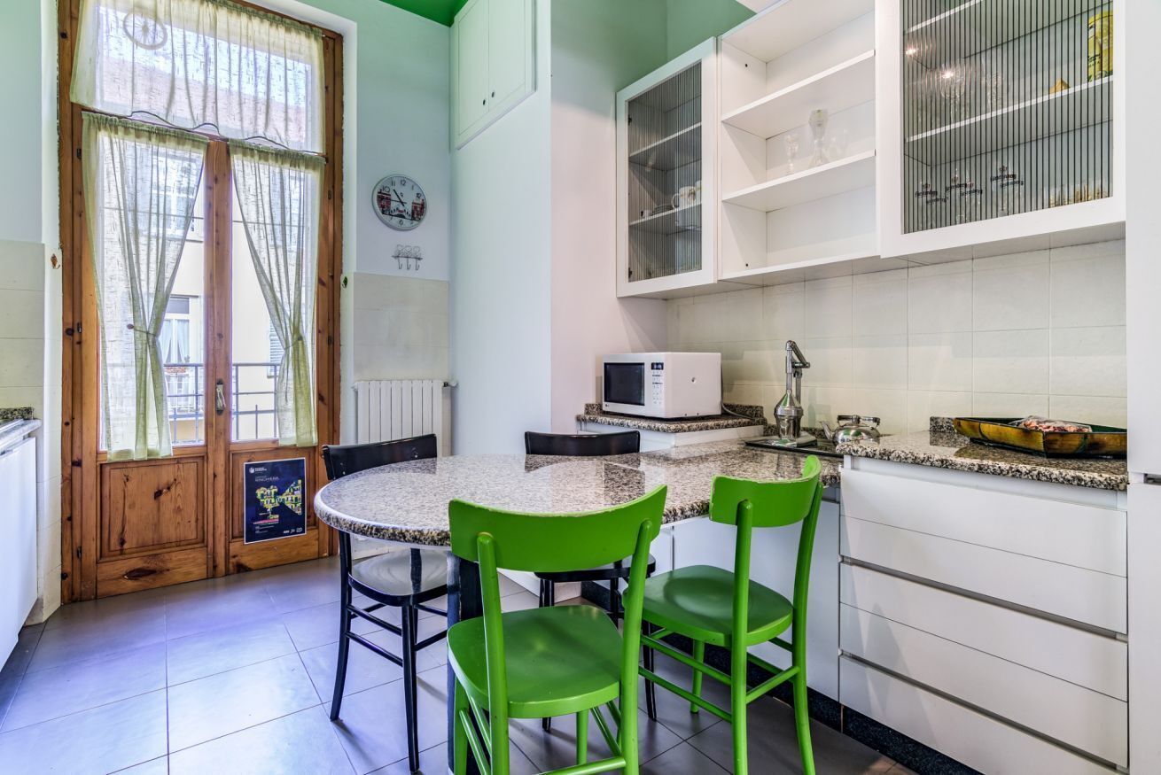A kitchen with a round table, two green chairs, a microwave, and a window with curtains.