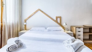 Ein ordentlich bezogenes Bett mit weißen Leinen, zwei Nachttischlampen, ein Holz-Kopfschild und ein Nachttisch mit Schublade.