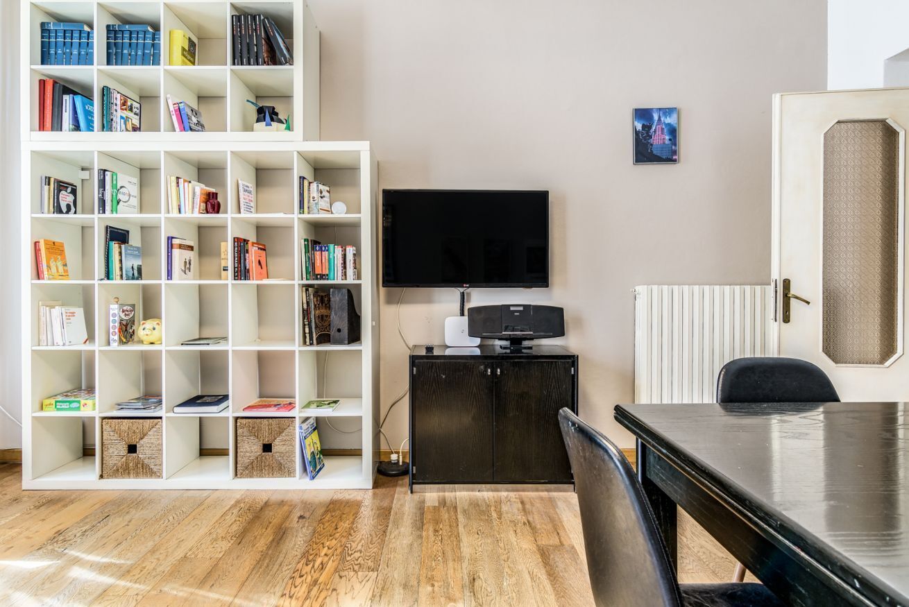 A room with a white bookshelf filled with books, a black TV on a cabinet, a wooden floor, and a black table with a chair.