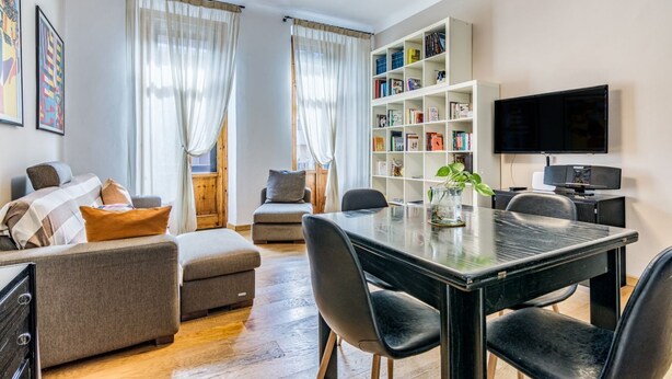 Ein modernes Wohnzimmer mit einer Couch, einem Hocker und einem Esstisch mit Stühlen. Ein Bücherregal und ein Flachbildfernseher an der Wand sind ebenfalls vorhanden.