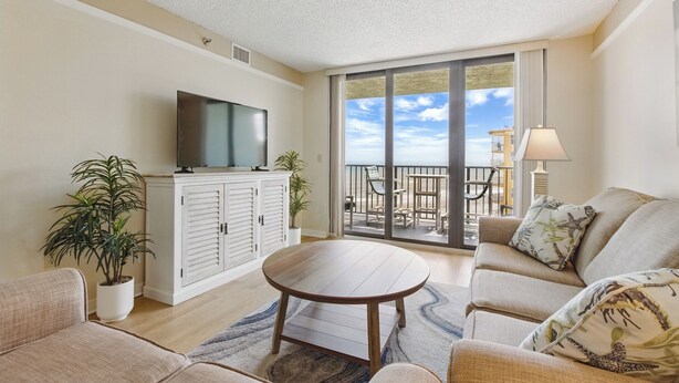 A living room with a flat-screen TV, a round wooden coffee table, a beige sofa, and a balcony with outdoor furniture. - Beachfront Bliss! Oceanfront Pool! Great Location!  Ocean Trillium 702 (New Smyrna Beach)