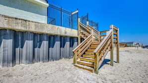 A wooden staircase leading to a balcony with a metal railing, a concrete wall, and a sandy beach in the background. - Beachfront Bliss! Oceanfront Pool! Great Location!  Ocean Trillium 702 (New Smyrna Beach)