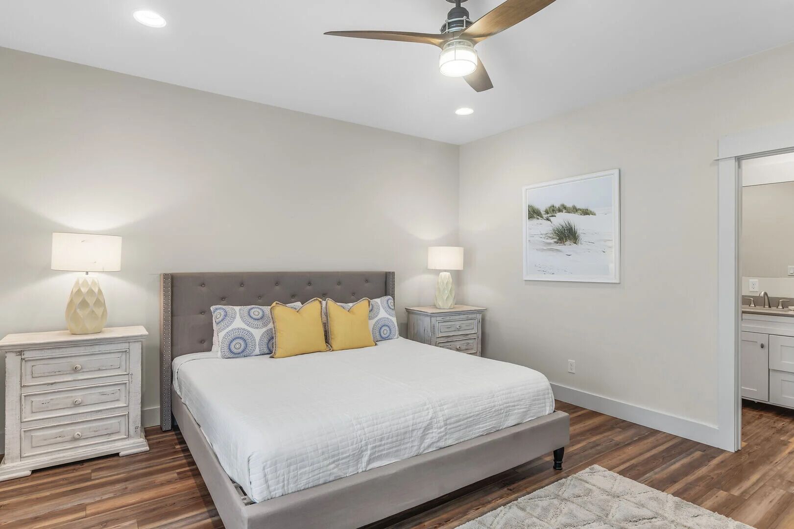 A bedroom with a bed, two bedside tables, a ceiling fan, a framed picture on the wall, and a bathroom visible through an open door.